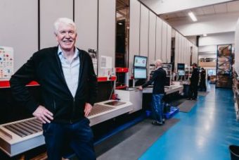 Daglig leder, Jan Kleven, smiler stolt til kamera foran Elotec sin AutoStore-løsning med ansatte som opererer portene i bakgrunnen.