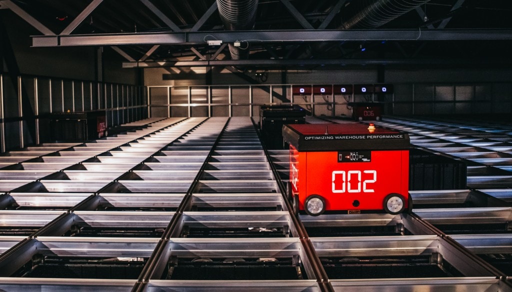 En rød AutoStore-robot kjører på aluminiumgriden høyt opp mot taket.